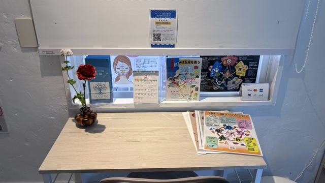 Display shelf with anime posters, magazines, and a red flower in a vase on wooden table